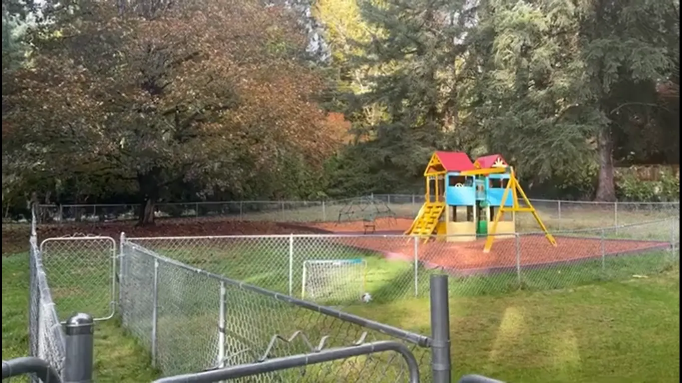 The fenced playground and play structure, surrounded by tall trees